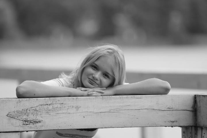Czarno-białe zdjęcie portretowe uśmiechniętej młodej blondynki opierającej się na pomalowanej na biało drewnianej balustradzie.