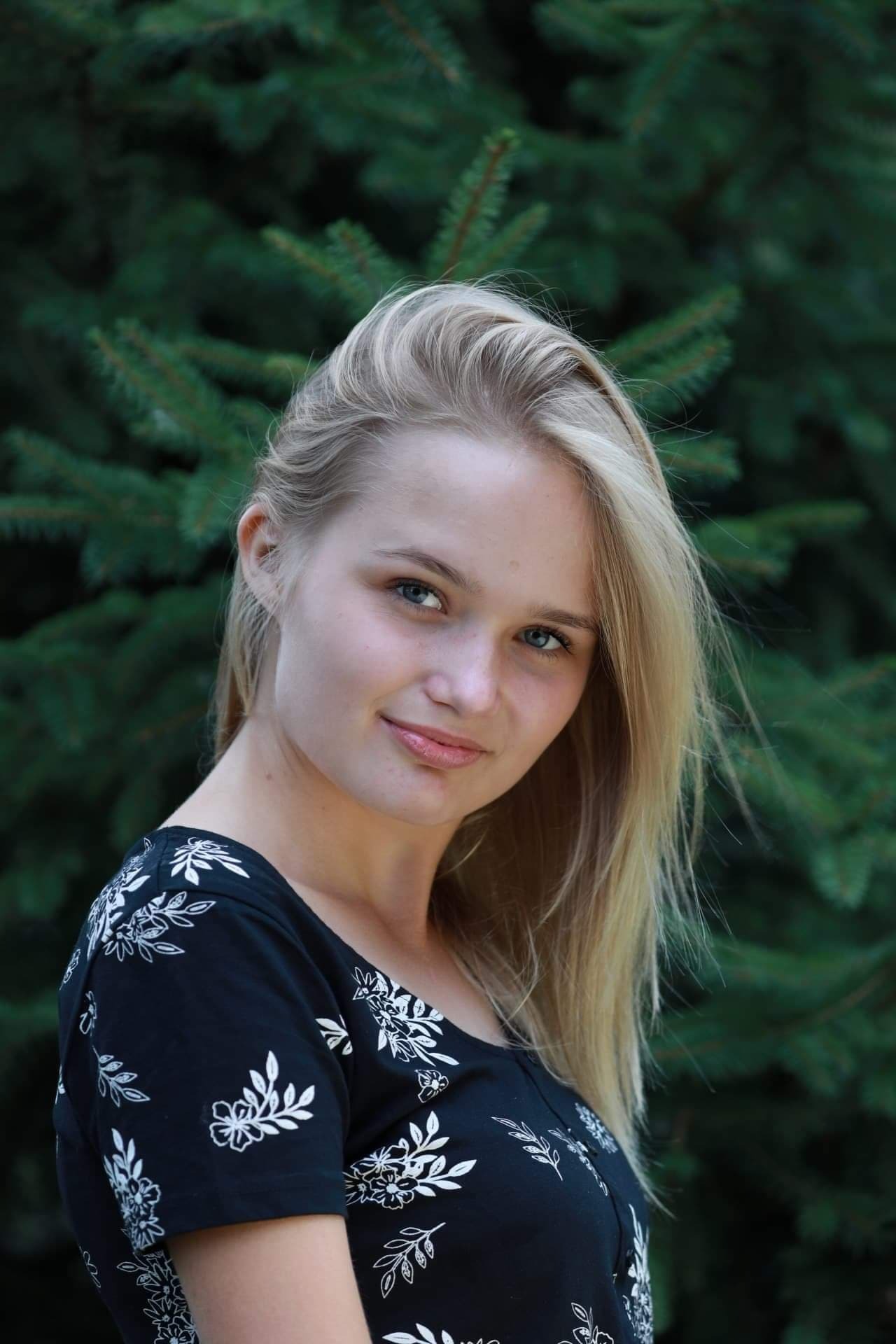 Portret młodej kobiety z blond włosami na tle iglastego drzewa, ubrana w czarną bluzkę w białe kwiaty, z delikatnym uśmiechem.