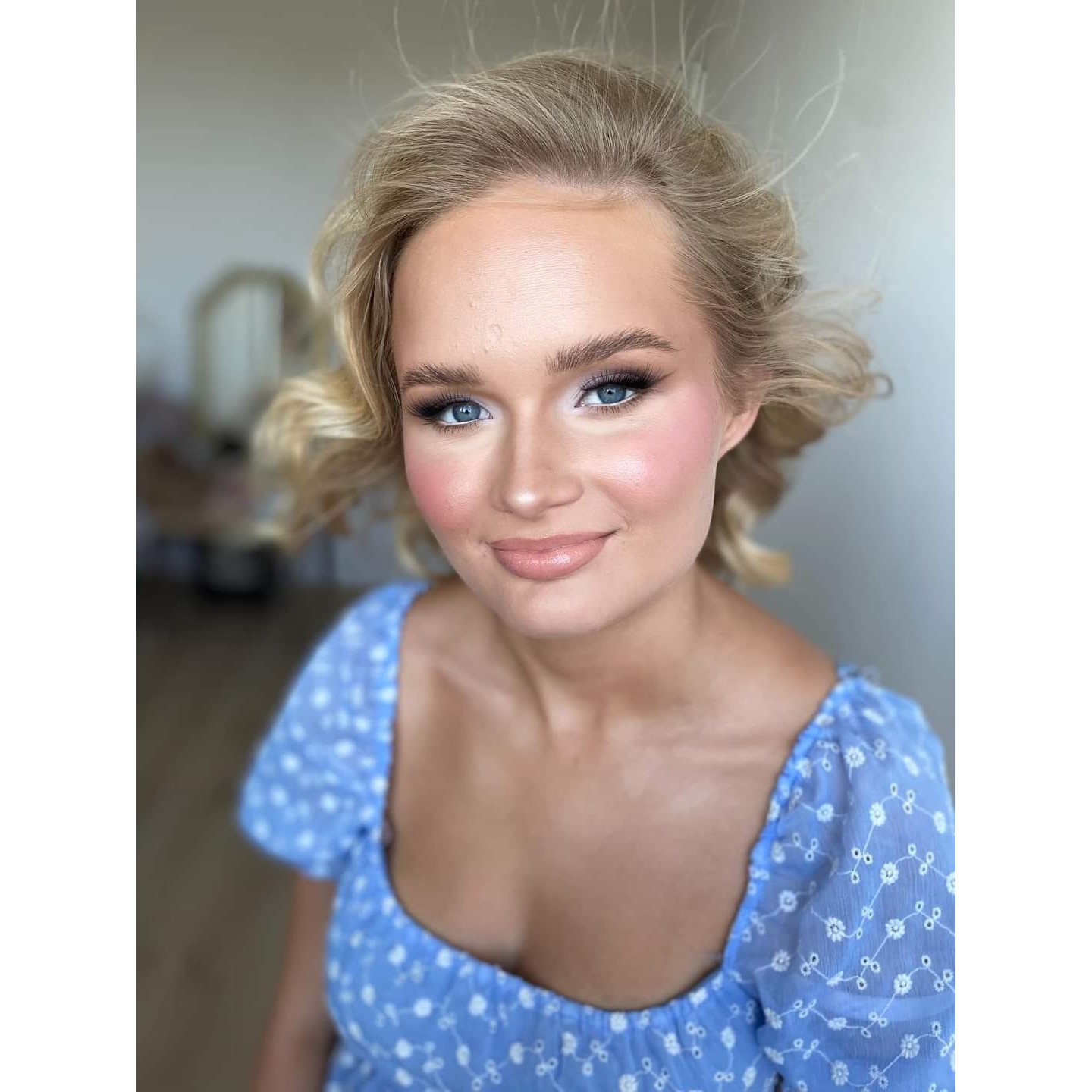 Portret uśmiechniętej blondynki z delikatnym makijażem i niebieskiej sukience w kwiatki, z widocznym odbiciem lustra w tle.