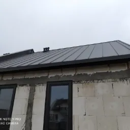 Szary dach z blachy na surowym budynku z widocznymi oknami i kominkami w tle pochmurne niebo.