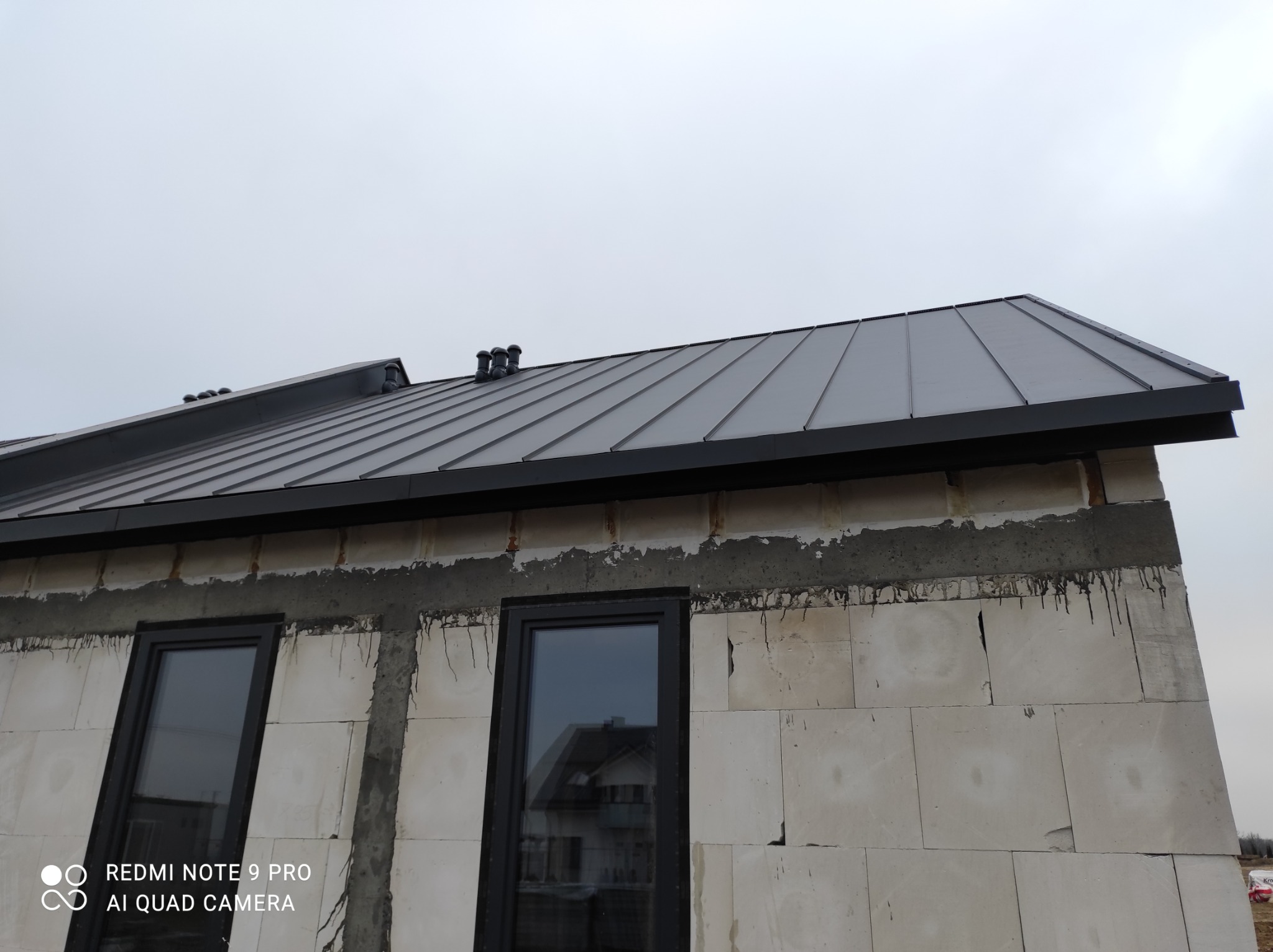 Szary dach z blachy na surowym budynku z widocznymi oknami i kominkami w tle pochmurne niebo.