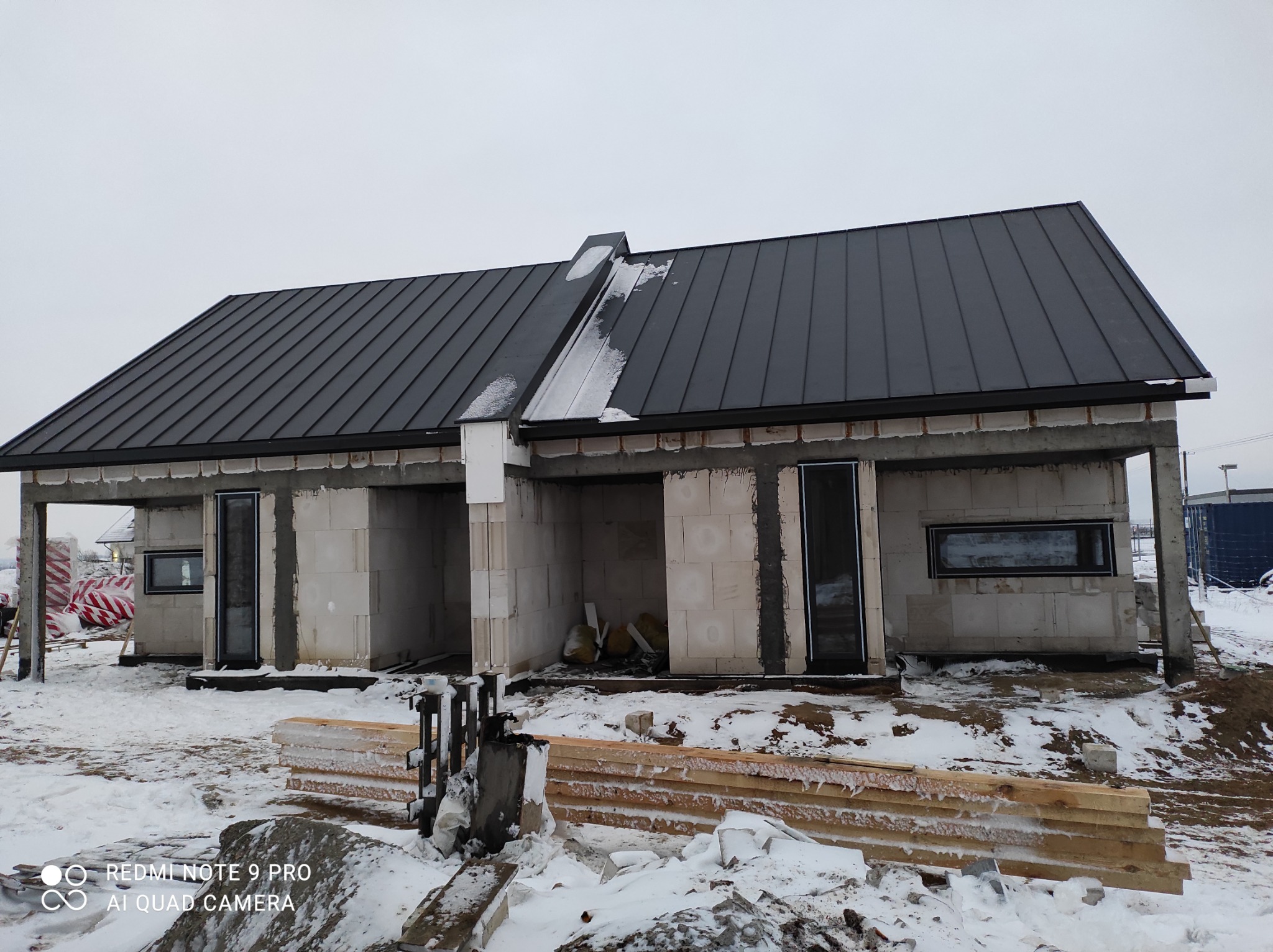 Surowy stan bliźniaka z ciemnoszarą blachodachówką na dachu, widoczne bloczki betonowe, częściowo ośnieżone, okna wstawione, na pierwszym planie stos drewna i śnieg.