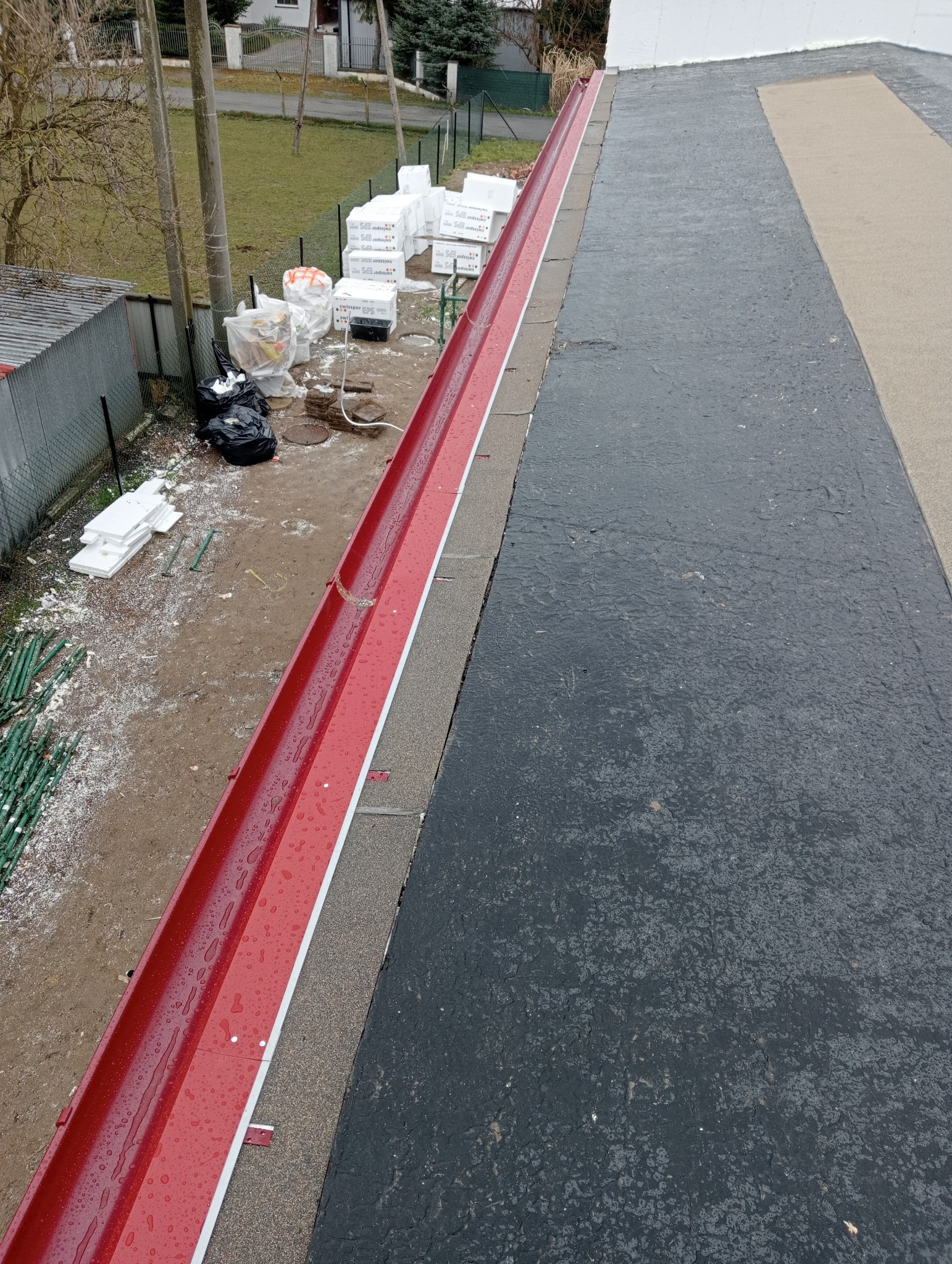 Nowa, błyszcząca, czerwona rynna zamontowana na dachu pokrytym papą, widoczne krople wody po deszczu, w tle materiały budowlane i teren posesji.