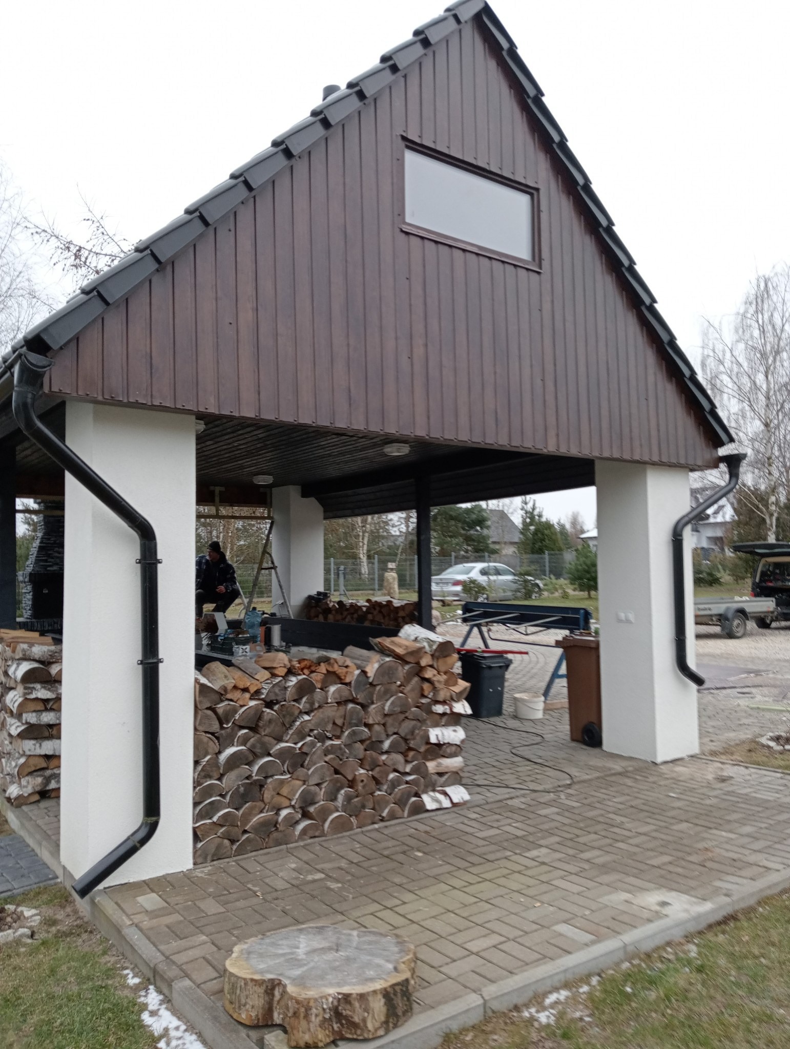 Zadaszona wiata z ciemnobrązową elewacją i czarnym dachem, widoczne rynny, stos drewna opałowego i osoba wewnątrz konstrukcji.