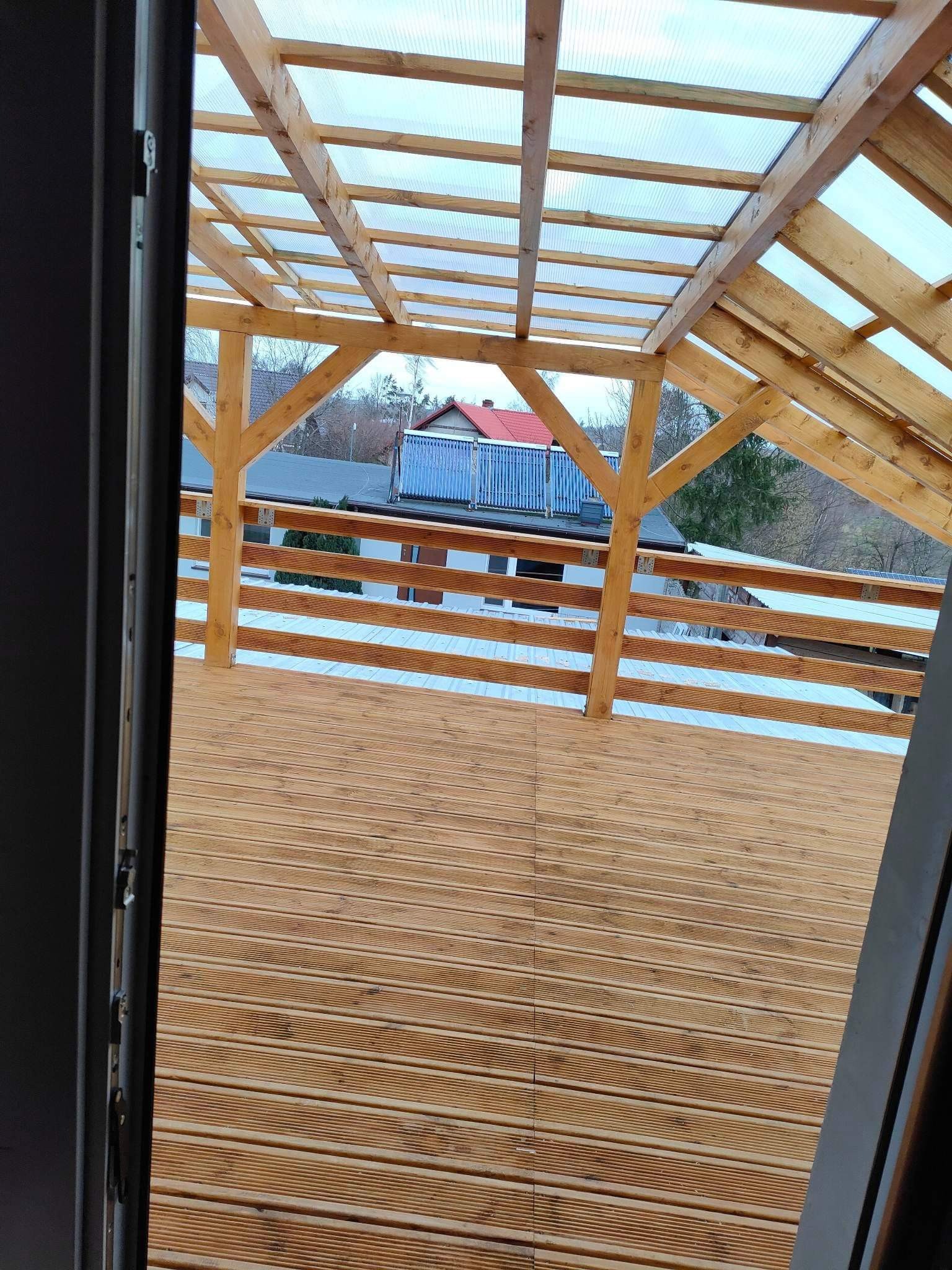 Drewniana konstrukcja dachu z przezroczystym pokryciem nad tarasem, widok z wnętrza przez otwarte drzwi balkonowe.