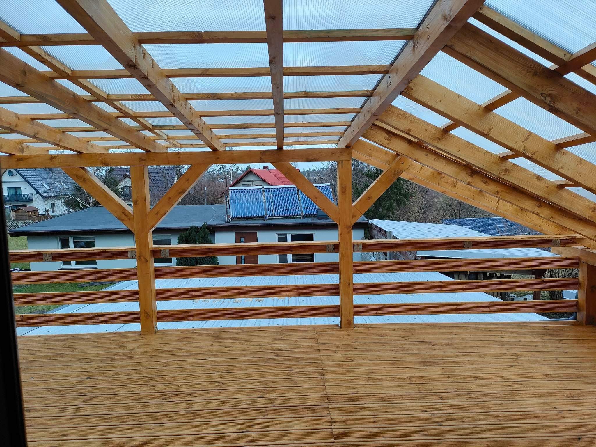 Drewniana konstrukcja z przezroczystym zadaszeniem na tarasie, widok z dołu na elementy konstrukcyjne i otoczenie zabudowane domami jednorodzinnymi.