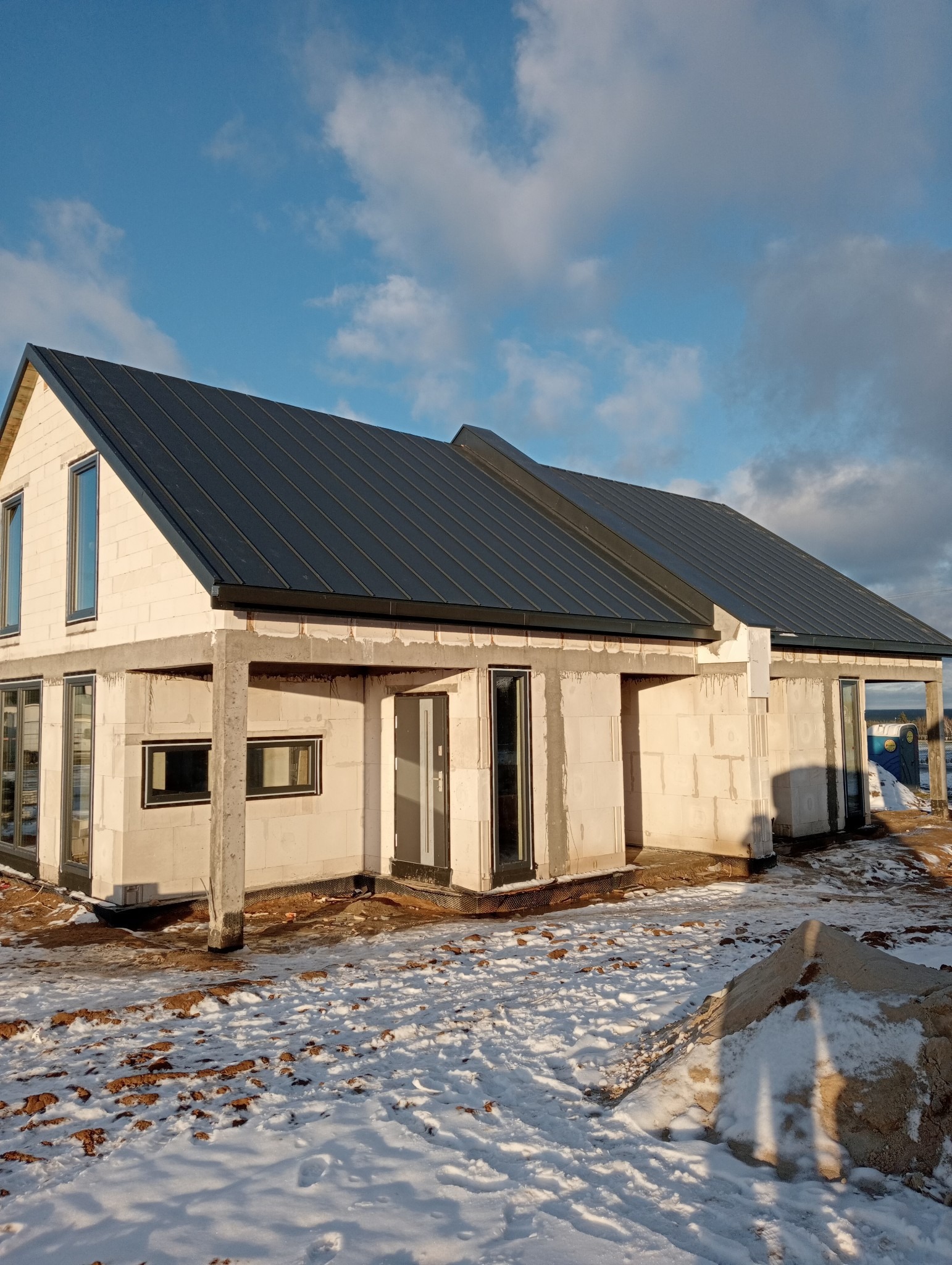 Surowy stan domu jednorodzinnego z ciemnoszarą blachą na dachu, widoczny w zimowej scenerii z resztkami śniegu na ziemi i stertą piasku. Dom w trakcie budowy, z widocznymi betonowymi elementami...