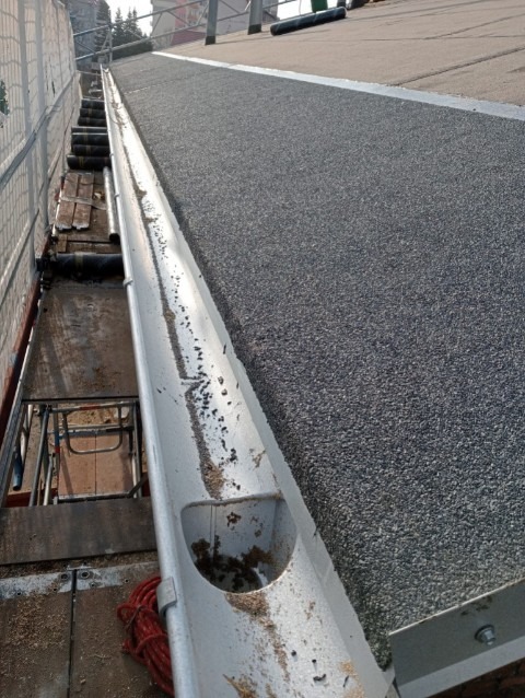 Widok z góry na fragment dachu pokrytego papą i zamontowaną, aluminiową rynną z widocznymi zabrudzeniami wewnątrz.