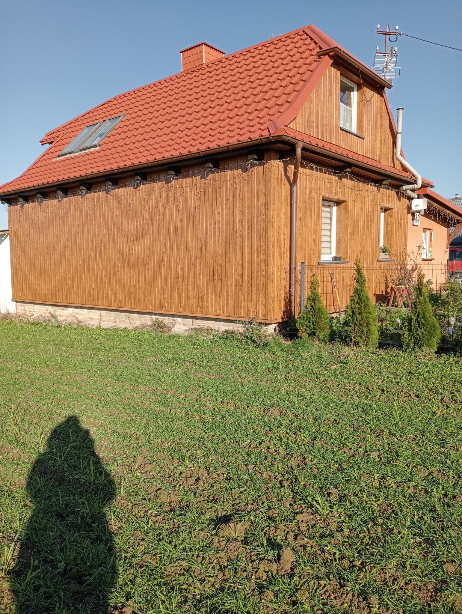 Dom jednorodzinny z czerwonym dachem z dachówki, drewnianą elewacją i widocznymi rynnami, oświetlony słońcem, z fragmentem trawnika na pierwszym planie i cieniem osoby.