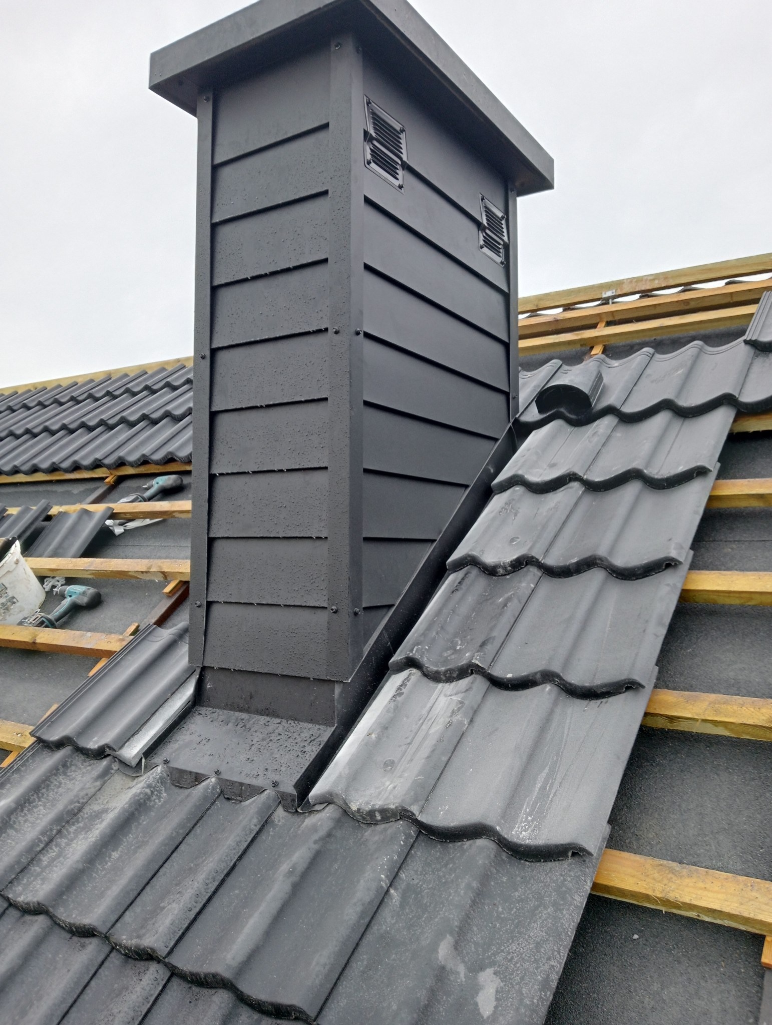 Czarny komin wentylacyjny z blachy trapezowej na dachu z dachówki ceramicznej w trakcie montażu, widoczne łaty drewniane i narzędzia.