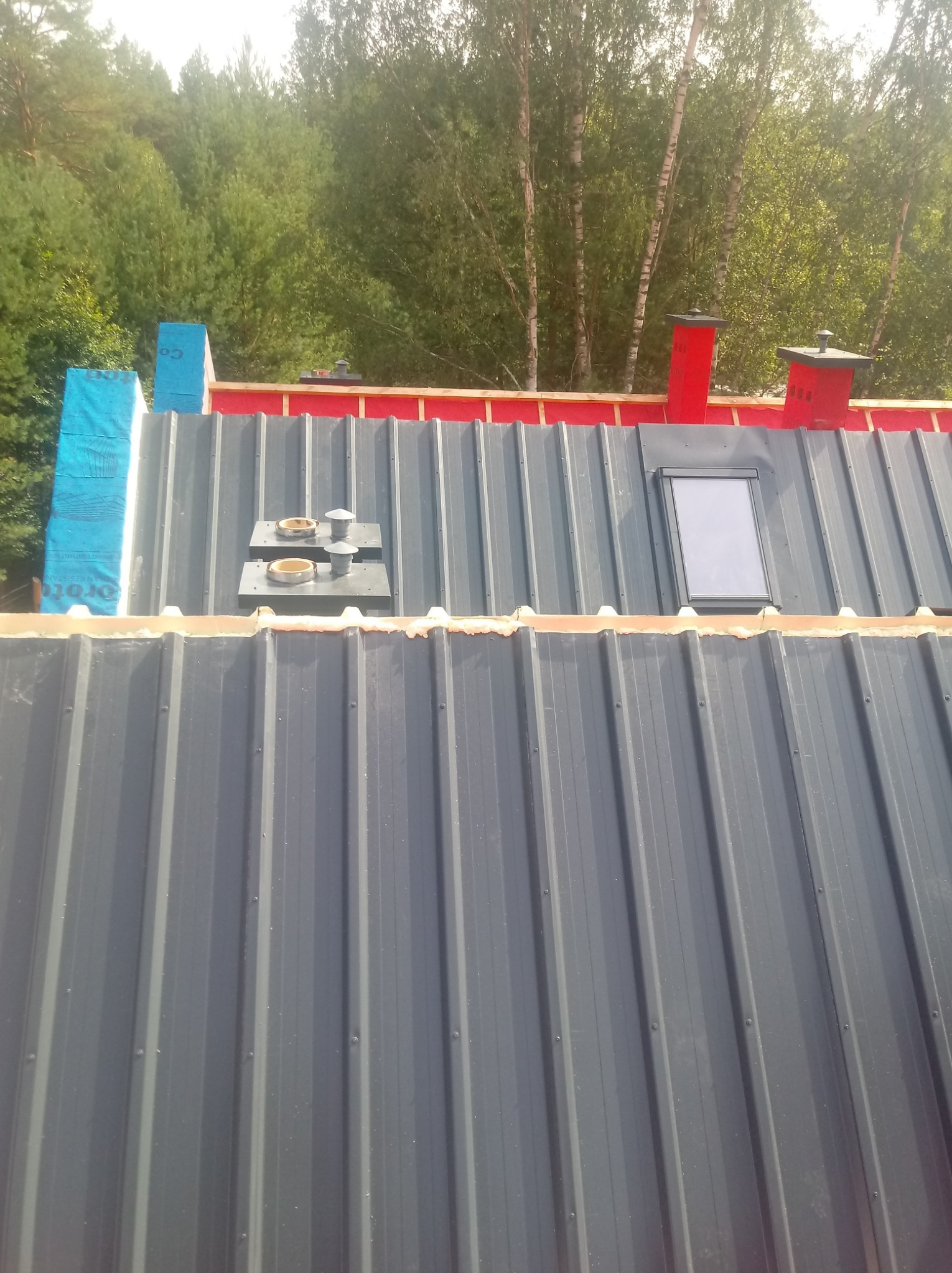 Szary dach z blachy trapezowej z oknem dachowym, kominkami wentylacyjnymi i obróbkami blacharskimi, widoczne zabezpieczenie folią paroizolacyjną na krawędzi dachu.