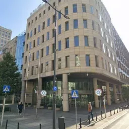 Elewacja biurowca z kamienną okładziną i szklanym wejściem, widok z poziomu ulicy, z widocznymi znakami drogowymi i przechodniami.