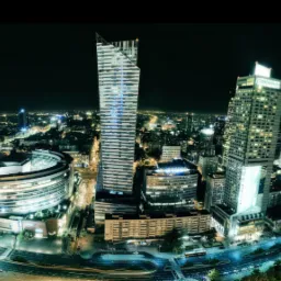 Nocna panorama Warszawy z widokiem na oświetlone drapacze chmur, w tym charakterystyczny budynek Warsaw Spire z ukośnym szczytem.