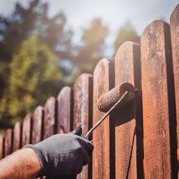 MasterfulMake - Malowanie drewnianego płotu brązową farbą za pomocą wałka, widoczna ręka w szarej rękawicy, w tle zieleń drzew.