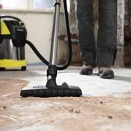 MasterfulMake - Odkurzacz przemysłowy z żółto-czarną obudową podczas pracy na betonowej podłodze w trakcie remontu, widoczne ślady po usuniętym parkiecie i fragmenty surowych ścian.