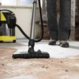 Odkurzacz przemysłowy z żółto-czarną obudową podczas pracy na betonowej podłodze w trakcie remontu, widoczne ślady po usuniętym parkiecie i fragmenty surowych ścian.