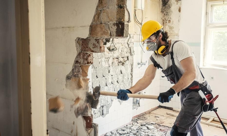 Pracownik w kasku i masce, z młotem w dłoni, wyburza fragment ściany z płytek w łazience, widoczne gruz i odsłonięta cegła.