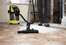 Odkurzacz przemysłowy z żółto-czarną obudową podczas pracy na betonowej podłodze w trakcie remontu, widoczne ślady po usuniętym parkiecie i fragmenty surowych ścian.