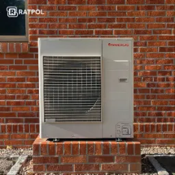 Biała pompa ciepła marki Immergas zamontowana na ceglanym cokole przy ceglanej ścianie budynku, widoczny fragment okna i logo firmy Ratpol w rogu.