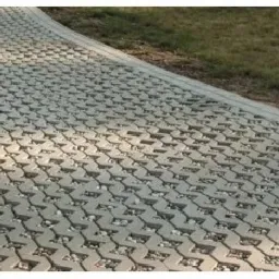 Szara, ażurowa nawierzchnia z kostki brukowej ekologicznej, wypełniona drobnym kamieniem, tworząca ścieżkę obok trawnika.