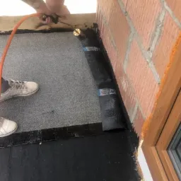 Prace dekarskie: Przyklejanie papy bitumicznej do krawędzi dachu przy ścianie z cegły za pomocą palnika gazowego. Widoczne białe tenisówki pracownika i butla z gazem.