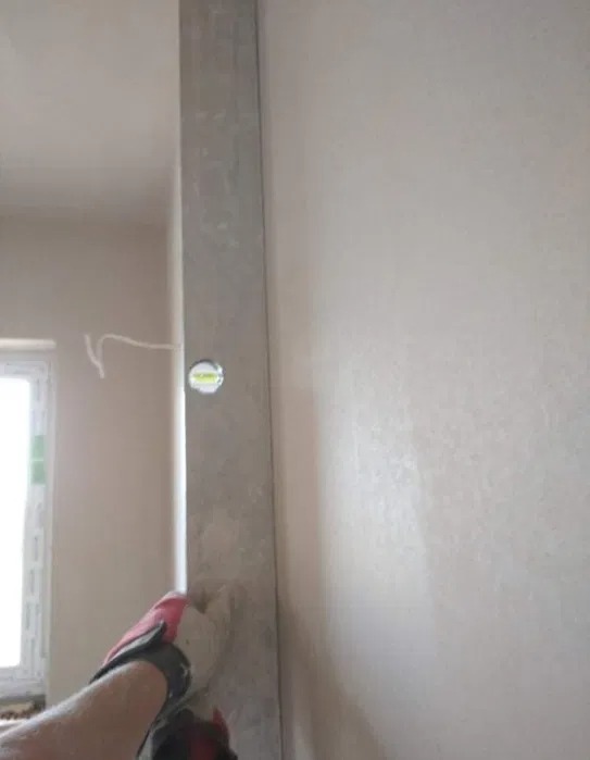 Nakładanie tynku maszynowego na narożnik ściany, widoczny poziomica i fragment okna w tle.