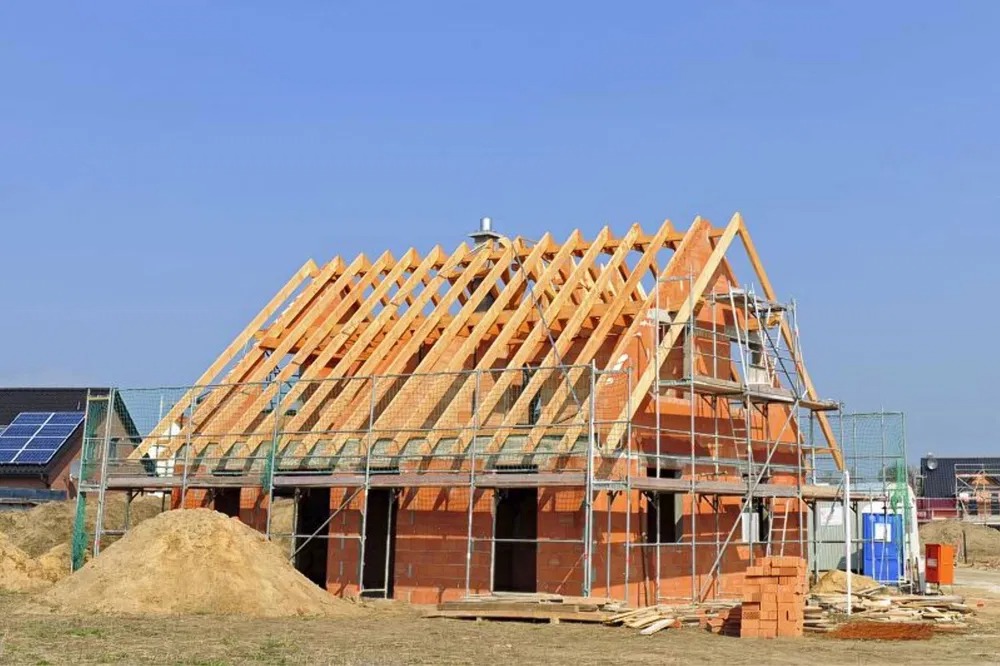 Budowa domu z czerwonej cegły: widoczne rusztowania, drewniana konstrukcja dachu w trakcie montażu, sterta piasku i materiały budowlane na placu budowy.