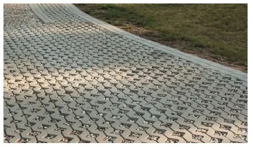 Szara, ażurowa nawierzchnia z kostki brukowej ekologicznej, wypełniona drobnym kamieniem, tworząca ścieżkę obok trawnika.