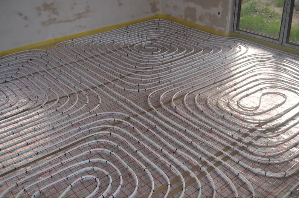 Rozłożona instalacja ogrzewania podłogowego z białych rur na folii z czerwoną kratką, widok z góry na fragment pomieszczenia z oknem.