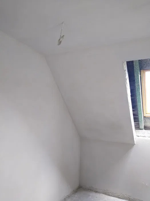 Wnętrze poddasza po nałożeniu białych tynków maszynowych, widoczne okno z zieloną folią izolacyjną i fragment zawieszonego kabla.