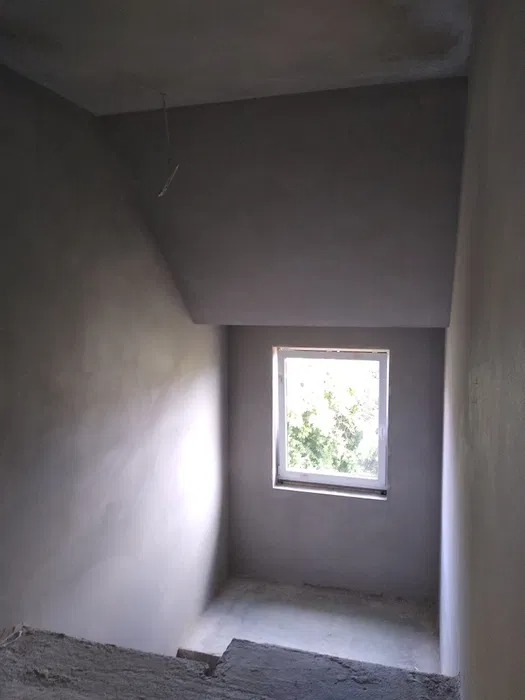 Wnętrze w trakcie prac wykończeniowych: świeżo otynkowane ściany w szarym odcieniu, widoczne niedoskonałości i fragmenty wystającej instalacji elektrycznej, małe okno z widokiem na zieleń na wprost.