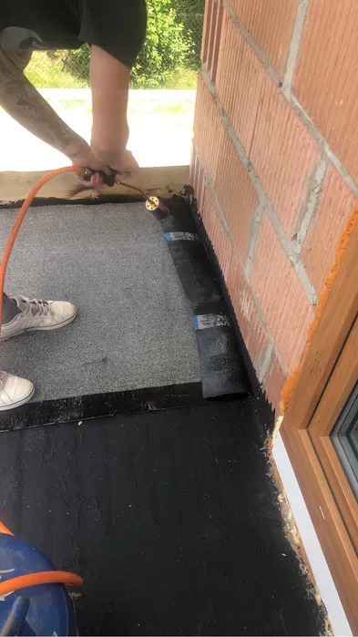 Prace dekarskie: Przyklejanie papy bitumicznej do krawędzi dachu przy ścianie z cegły za pomocą palnika gazowego. Widoczne białe tenisówki pracownika i butla z gazem.