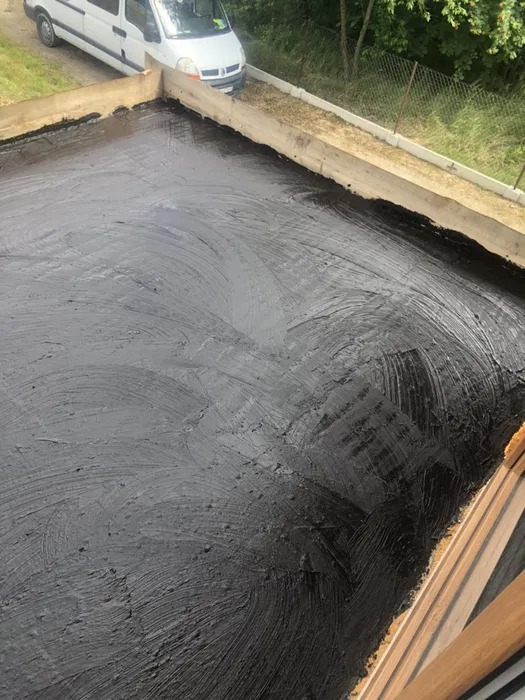 Czarna, świeżo nałożona hydroizolacja na płaskim dachu z drewnianym obramowaniem, widoczny biały samochód dostawczy w tle.
