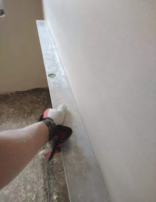 Nakładanie tynku na ścianę z użyciem długiej poziomicy; widoczna ręka w roboczej rękawicy, ściana pokryta świeżym tynkiem, surowa podłoga.