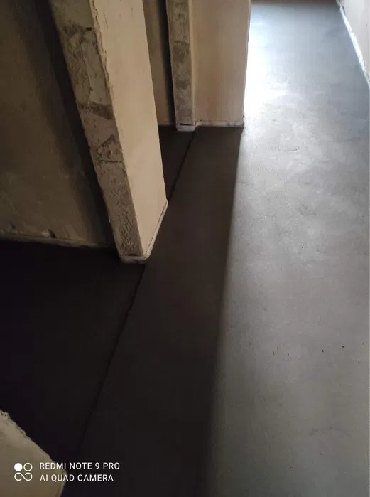 Świeżo wylana, gładka posadzka jastrychowa wewnątrz budynku, widoczne surowe ściany i progi drzwiowe, różnica w odcieniach szarości sugeruje etap schnięcia.