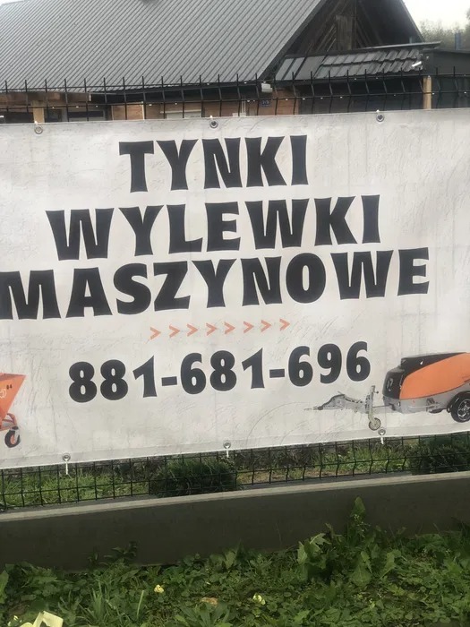 Baner reklamowy z napisem 'Tynki Wylewki Maszynowe' oraz numerem telefonu, umieszczony na ogrodzeniu z siatki, z widoczną przyczepą z maszyną do wylewek i taczką.