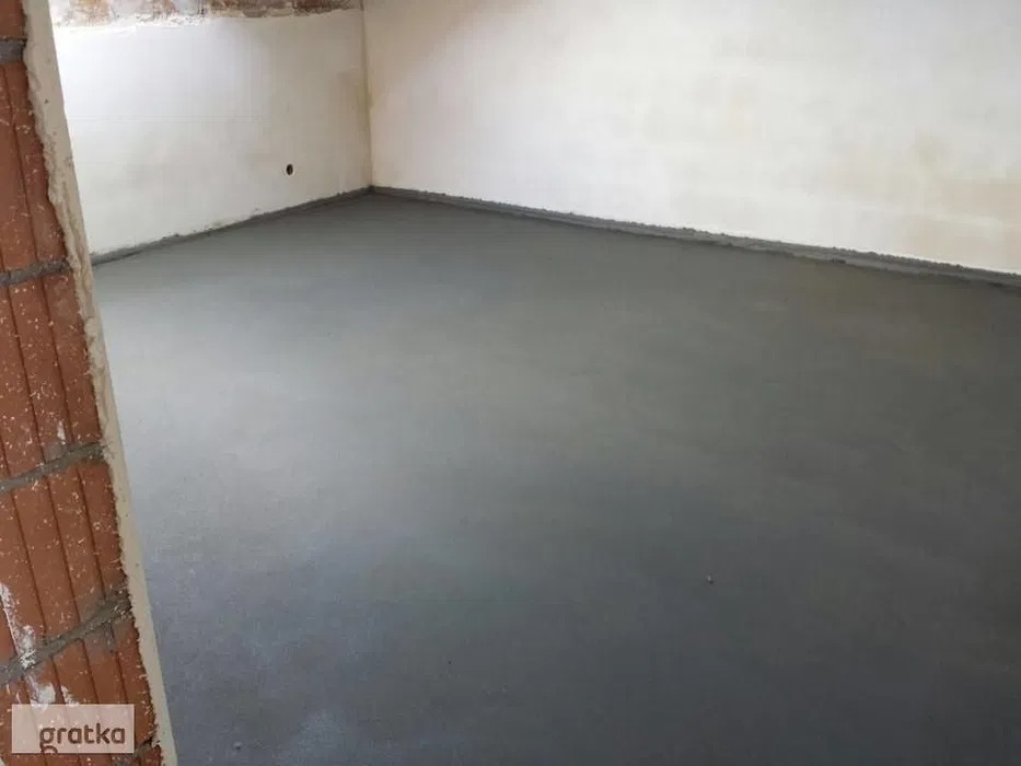 Świeżo wylana, gładka, szara wylewka jastrychowa w pomieszczeniu z surowymi ścianami. Fragment muru z czerwonej cegły w lewym dolnym rogu.