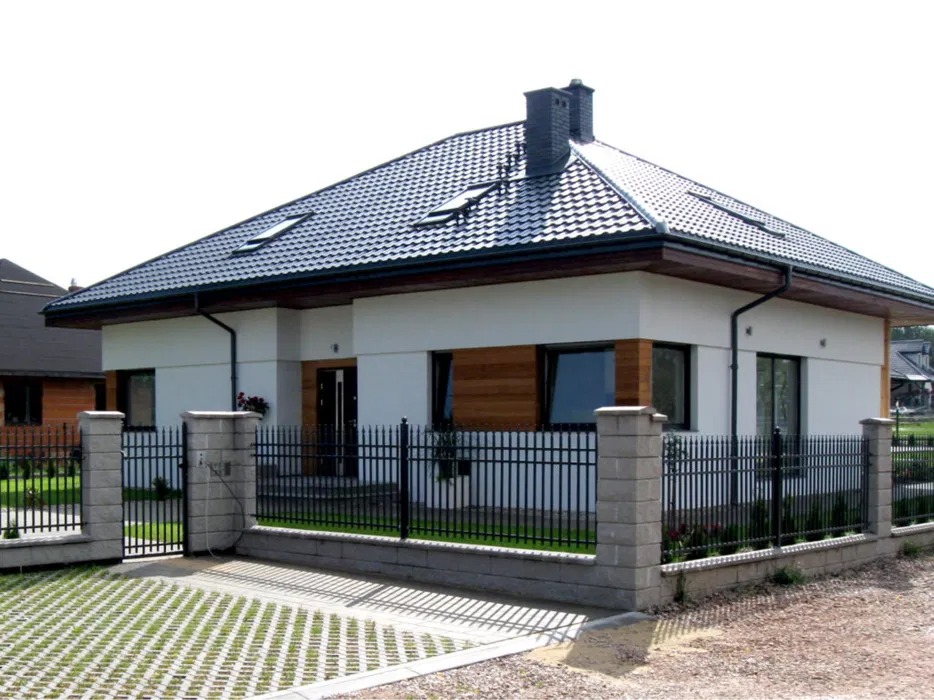 Nowoczesny dom jednorodzinny z czarnym dachem, oknami dachowymi i elementami drewnianej elewacji, ogrodzony czarnym metalowym płotem z betonowymi słupkami, z ażurowym podjazdem z kostki betonowej.