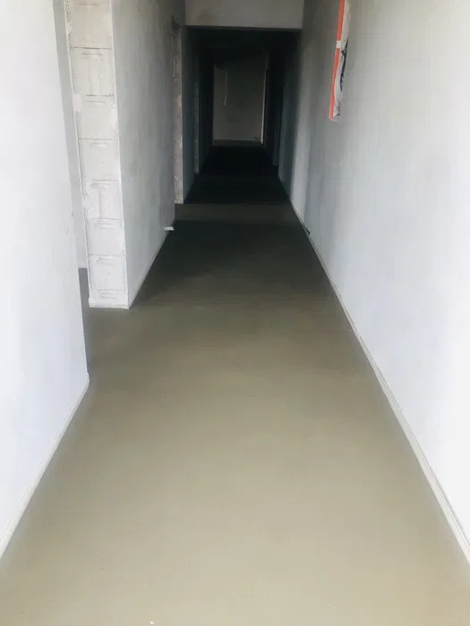Wnętrze w trakcie prac wykończeniowych: świeżo wylana, gładka posadzka w długim korytarzu z surowymi ścianami i widocznymi bloczkami. W tle widoczne ciemne przejście do kolejnych pomieszczeń.