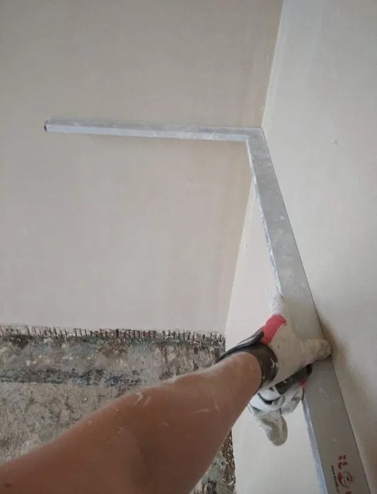 Ręka w rękawicy wyrównuje świeżo nałożony tynk przy użyciu aluminiowej łaty w rogu pomieszczenia. W tle widoczny fragment surowej ściany przygotowanej do tynkowania.