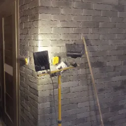 Ściana z dekoracyjnej cegły w trakcie montażu, oświetlona lampami budowlanymi na żółtym statywie, widoczne gniazdka elektryczne i fragment osoby w jeansach.