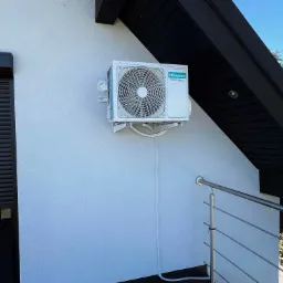 Biała jednostka zewnętrzna klimatyzacji Hisense zamontowana na ścianie budynku z czarnymi elementami architektonicznymi i fragmentem balustrady.