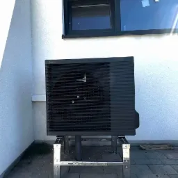 Czarna pompa ciepła Panasonic na metalowej konstrukcji przed białą ścianą z oknem. Widoczne detale montażu i otoczenie budynku.