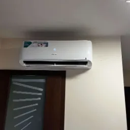 Biała klimatyzacja Hisense zamontowana nad ciemnymi drzwiami z geometrycznym wzorem na jasnej ścianie. Widoczny wyświetlacz temperatury.