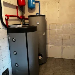 Dwa szare zbiorniki na wodę podłączone do instalacji z czerwonymi i niebieskimi elementami, widoczne płytki na ścianie i podłodze, oraz biała szafka w tle.