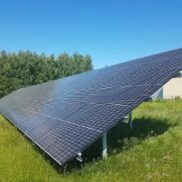 Instalacja fotowoltaiczna w Ujeździe, panele na metalowej konstrukcji, otoczone wysoką trawą, w tle budynek i drzewa pod błękitnym niebem.