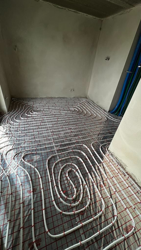 Białe rury ogrzewania podłogowego ułożone na folii z czerwoną siatką w surowym pomieszczeniu, widoczne niebieskie rury w rogu.