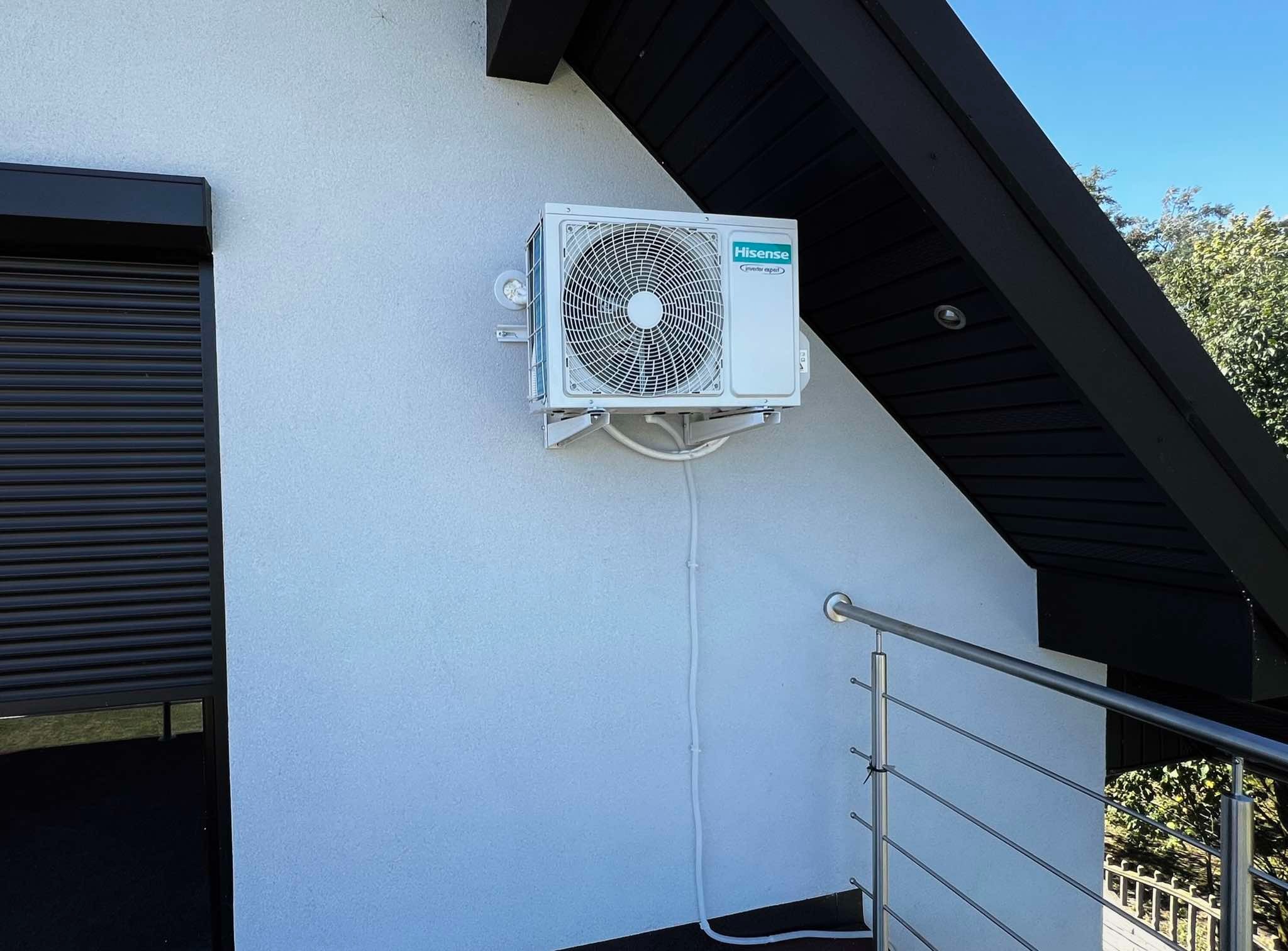 Biała jednostka zewnętrzna klimatyzacji Hisense zamontowana na ścianie budynku z czarnymi elementami architektonicznymi i fragmentem balustrady.