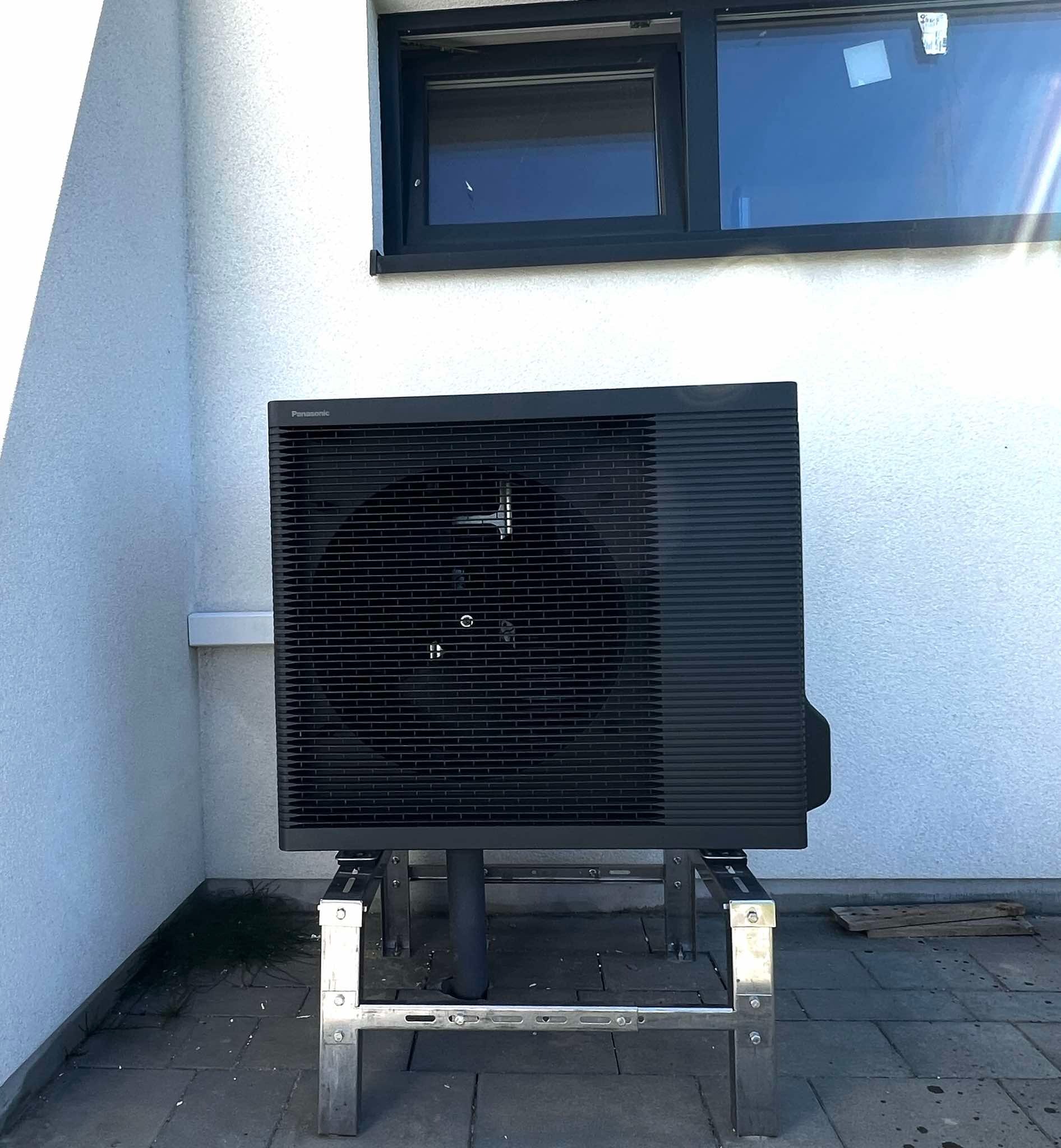 Czarna pompa ciepła Panasonic na metalowej konstrukcji przed białą ścianą z oknem. Widoczne detale montażu i otoczenie budynku.