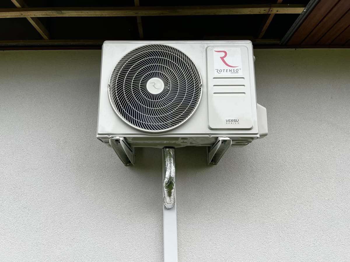 Biała jednostka zewnętrzna klimatyzacji Rotenso Versu zamontowana na jasnej ścianie budynku z widocznymi elementami instalacji.